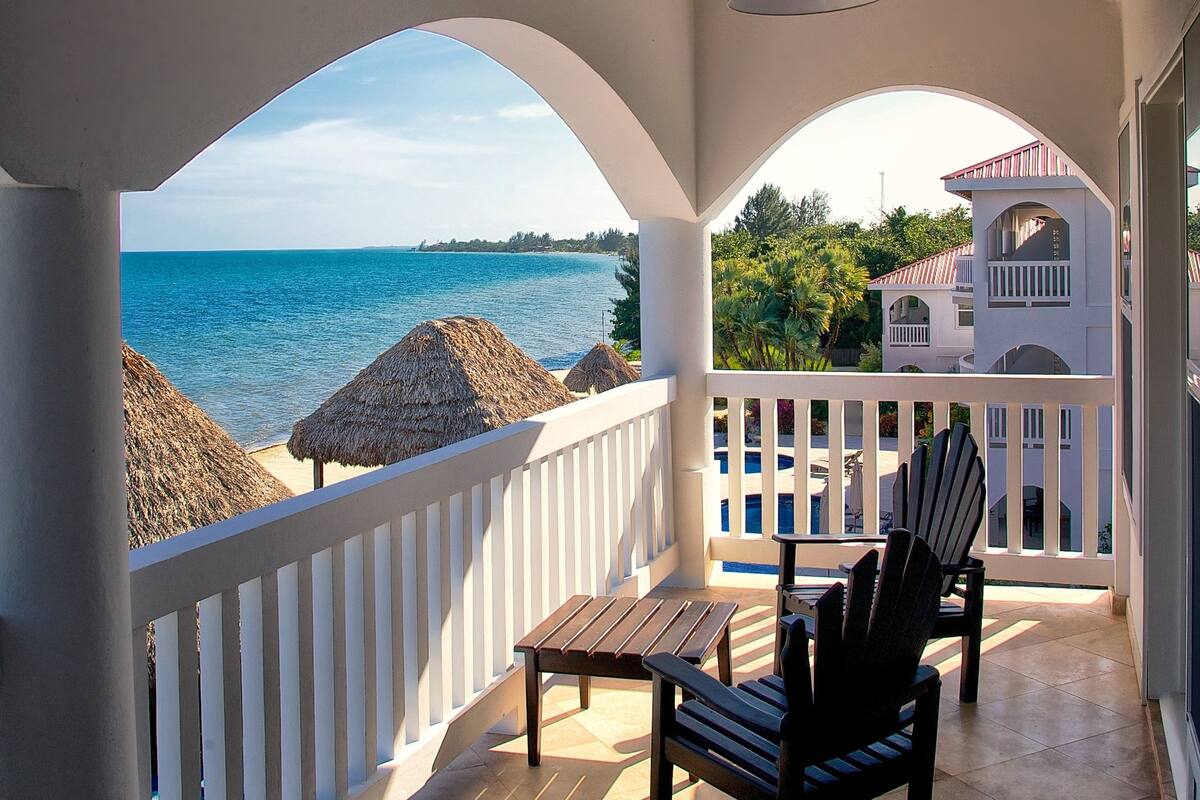 A covered balcony with a view of the sea, a pool, and a building in the background.