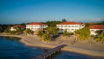 Un hotel frente a la playa con varias construcciones, un muelle de madera y palmeras.