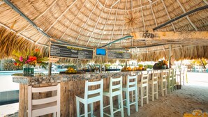 A beach bar with a thatched roof, white bar stools, and a menu board. - Umaya Resort & Adventures (Placencia)
