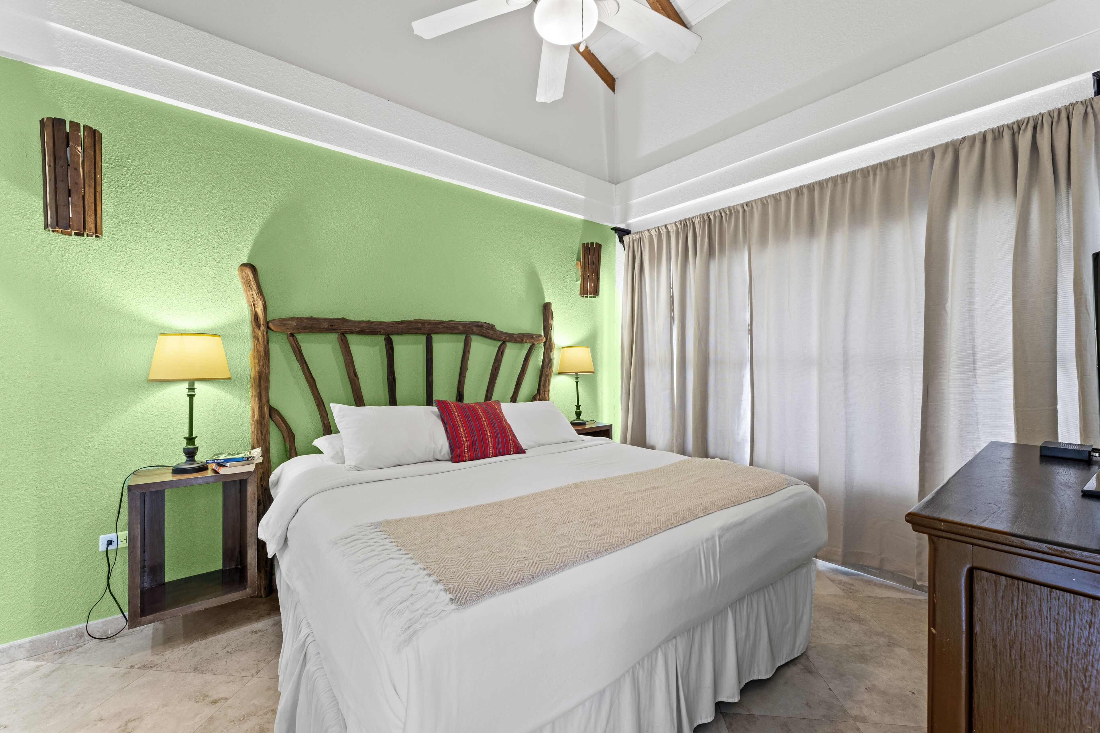 A neatly made bed with white bedding and a beige throw, a wooden headboard, a nightstand with a lamp, and a window with curtains.