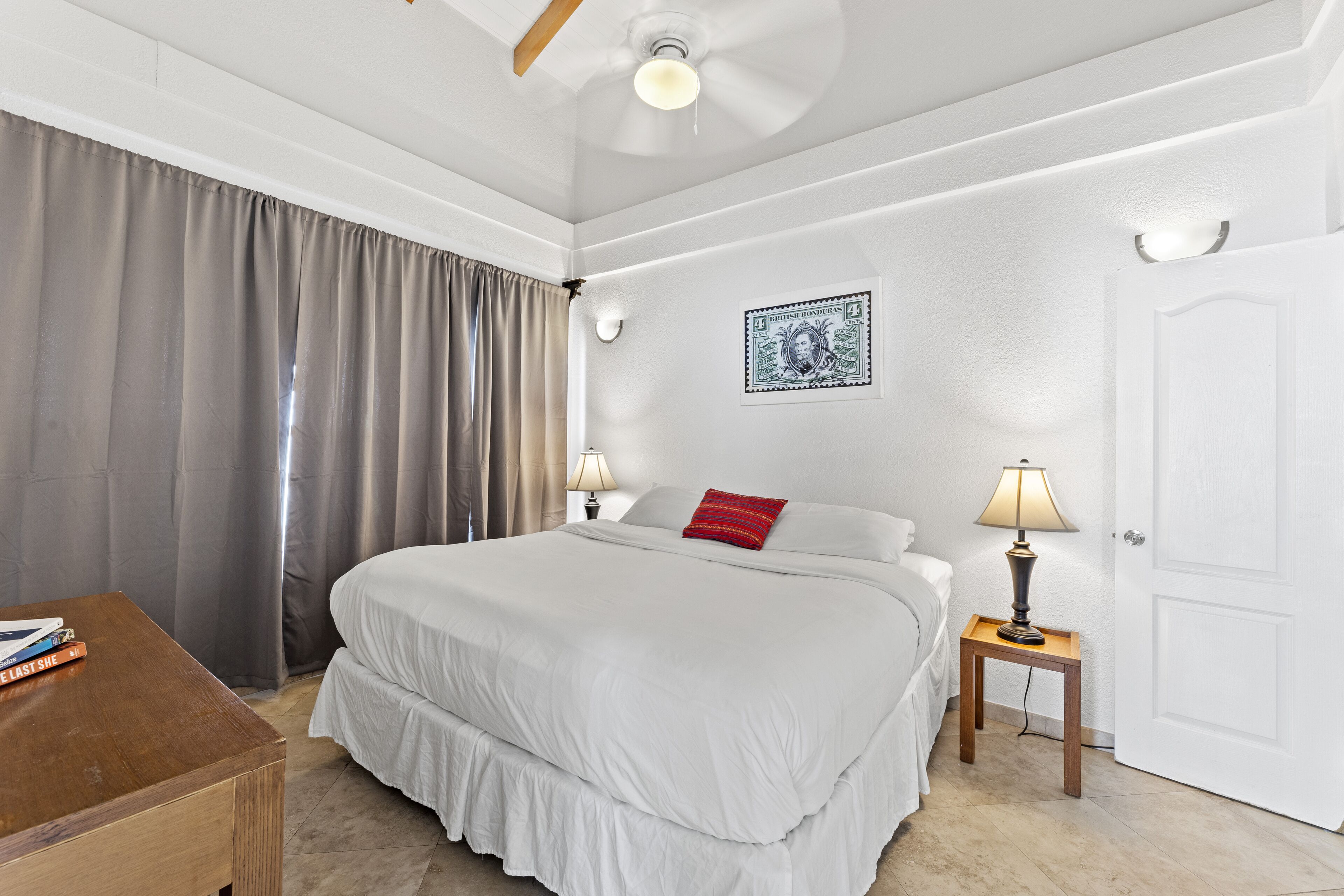 Habitación de hotel con una cama grande, dos mesitas de noche con lámparas, un ventilador de techo, una puerta y una ventana con cortinas.
