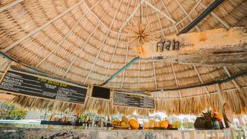 A beach bar with a thatched roof, wooden counter, and a menu board listing drinks and food items.