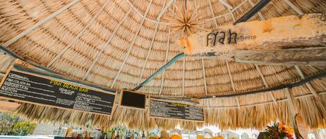 A beach bar with a thatched roof, wooden counter, and a menu board listing drinks and food items.