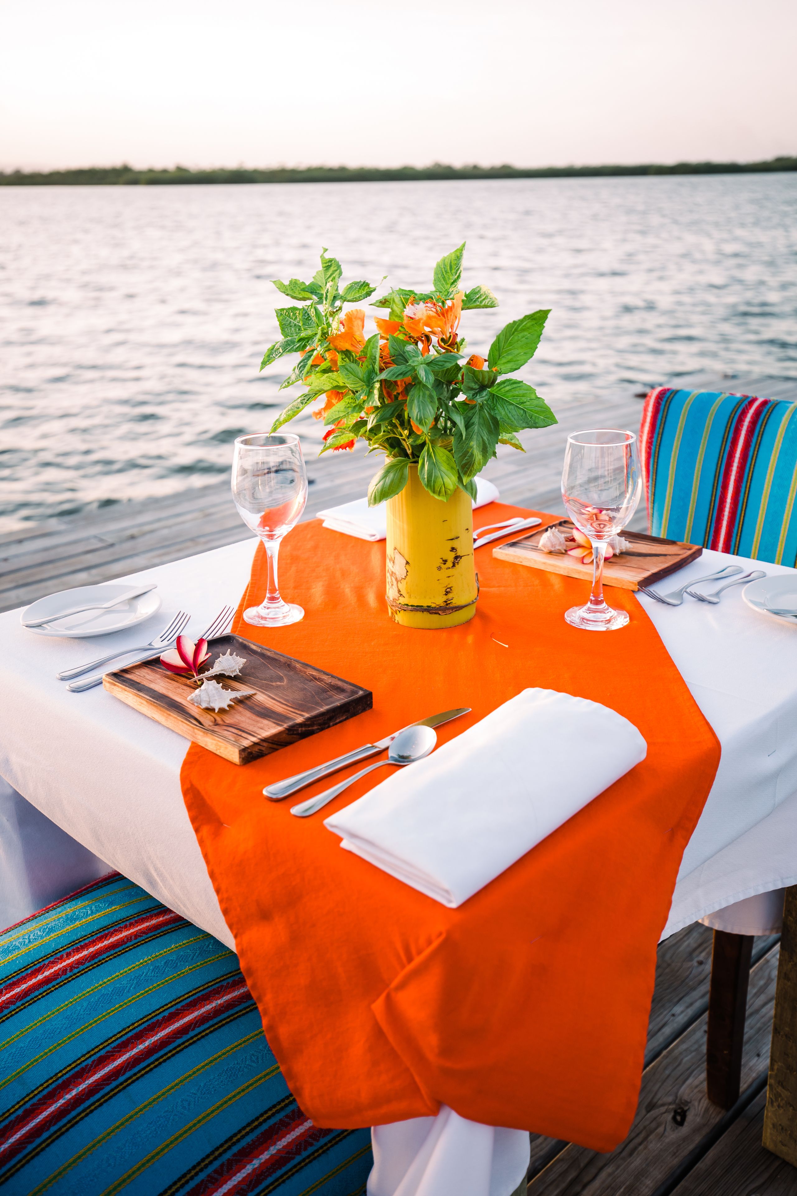 A table set for two with a yellow vase, two wine glasses, and a wooden board with food.