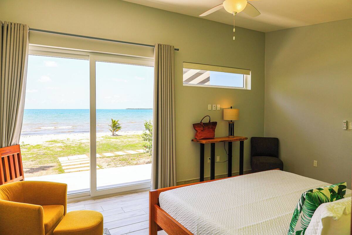 A hotel room with a bed, a chair, a table with a lamp, a handbag, and a view of the beach and ocean through a sliding door.