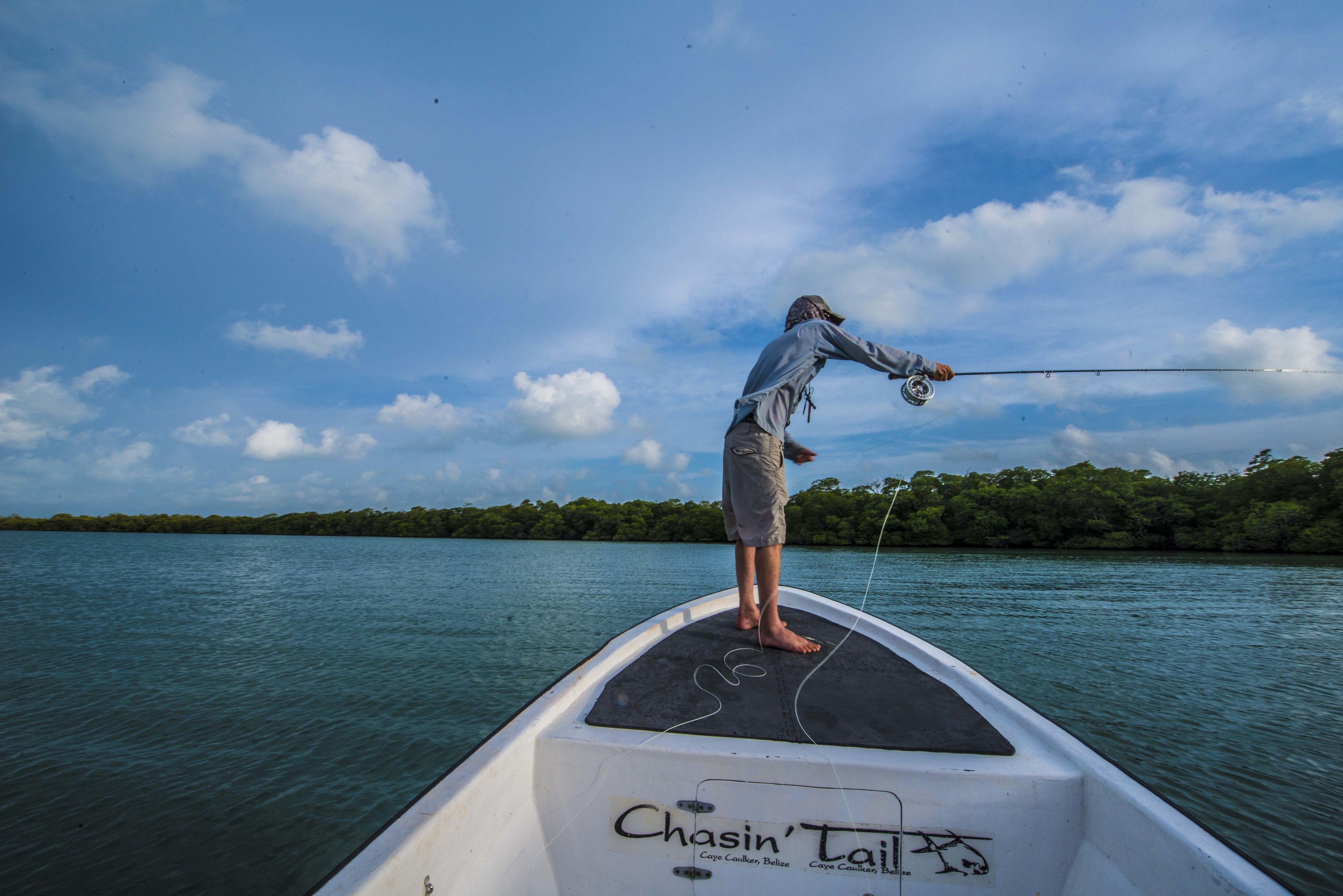 A person fishing from a boat named 'Chasin' Tail' with a calm water body and a forested shoreline in the background.