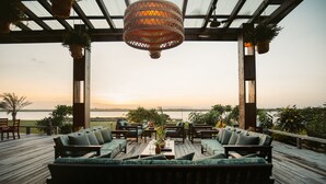 Terraza exterior con mobiliario de madera y plantas colgantes, con vista a una masa de agua durante el atardecer.