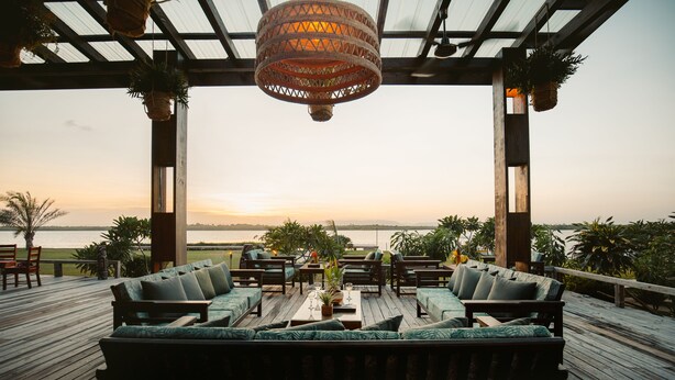 An outdoor deck with wooden furniture and hanging plants, overlooking a body of water during sunset. - Umaya Resort & Adventures (Placencia)