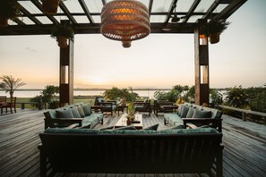 An outdoor deck with wooden furniture and hanging plants, overlooking a body of water during sunset. - Umaya Resort & Adventures (Placencia)