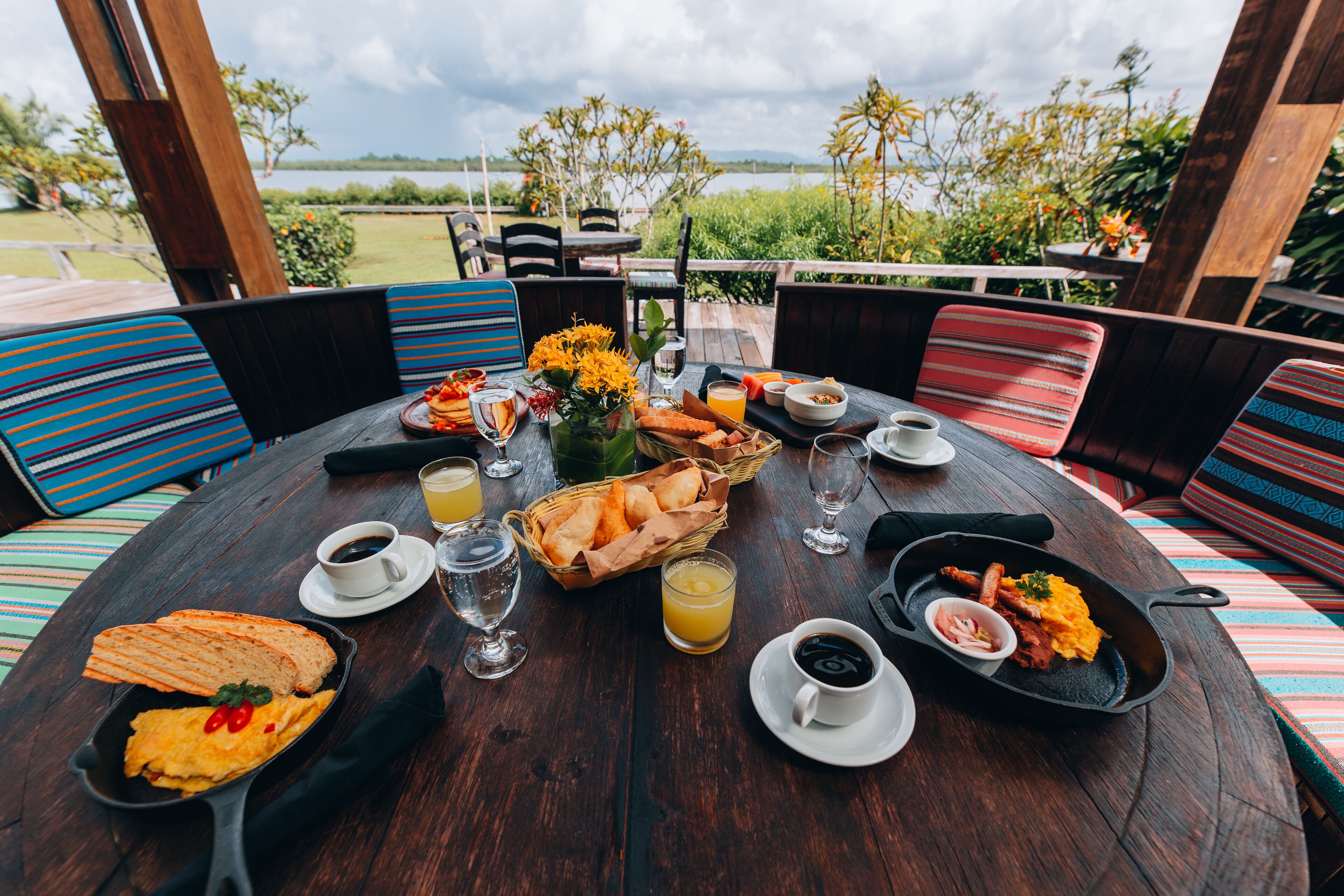 Una mesa de madera para comer con alimentos para el desayuno, incluyendo huevos, pan y bebidas, ubicada al aire libre con vista a un lago y vegetación.