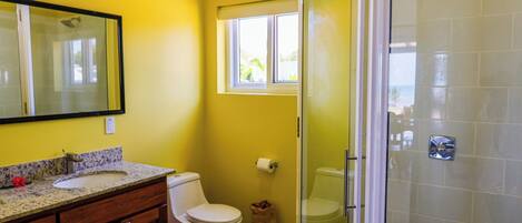A bathroom with a yellow wall, a white toilet, a sink with a granite countertop, and a glass-enclosed shower area.