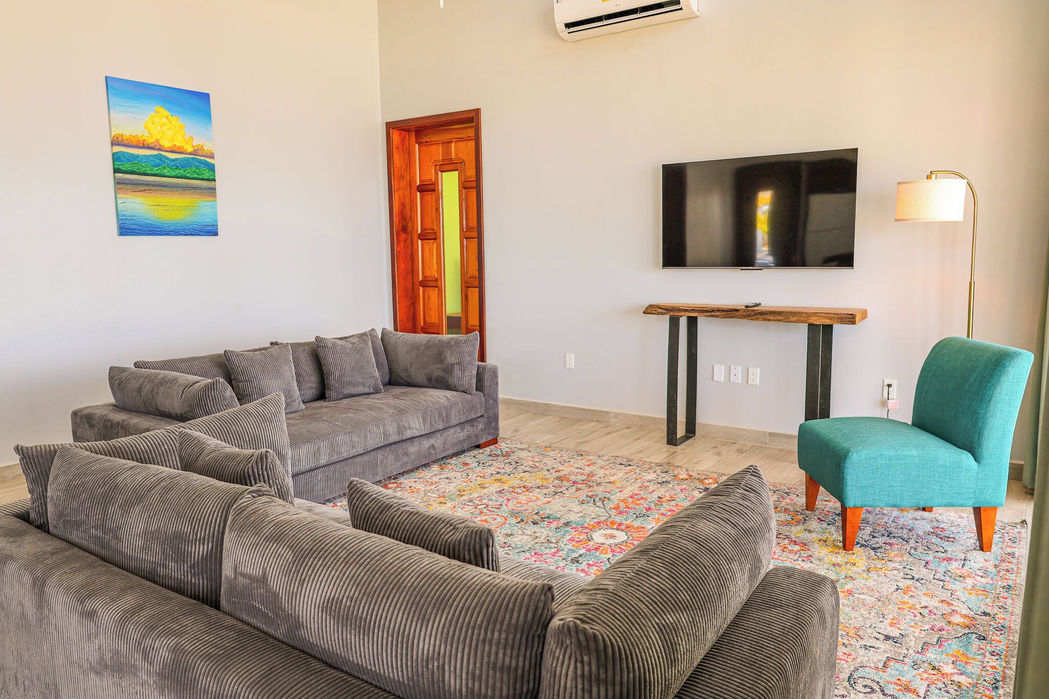 A living room with a grey sectional sofa, a teal armchair, a colorful rug, a wooden coffee table, a TV on the wall, and a door with a wooden frame.