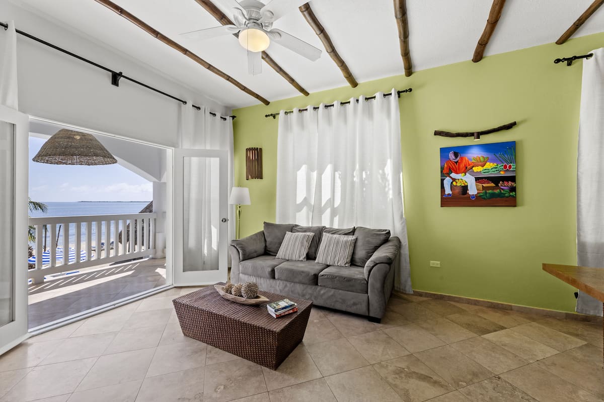 A living room with a grey sofa, a wicker coffee table, and a view of the beach through a glass door.