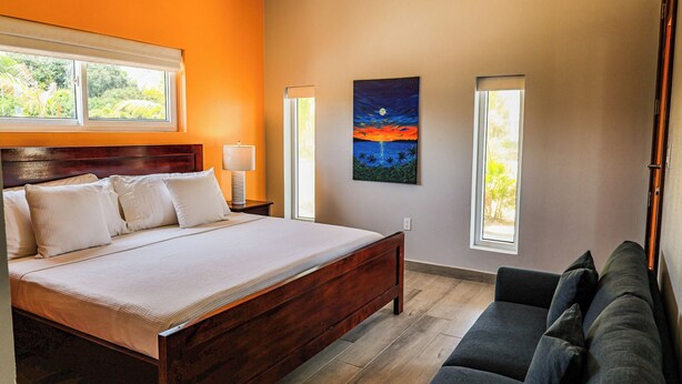 Un dormitorio con una cama, una mesita de noche, un cuadro y dos ventanas.
