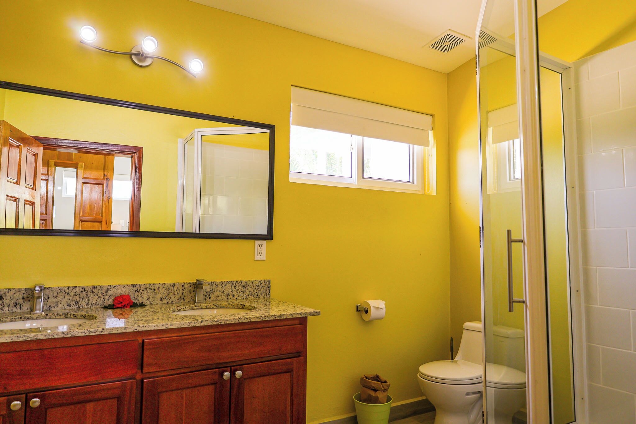 Une salle de bain avec des murs jaunes, un comptoir en granit, un meuble-lavabo en bois, des toilettes blanches et une douche à parois de verre.