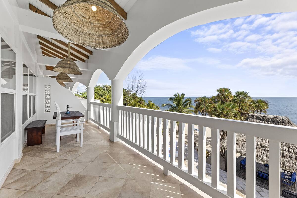 A covered balcony with a white railing, a table, and chairs, overlooking a beach with palm trees and the ocean.