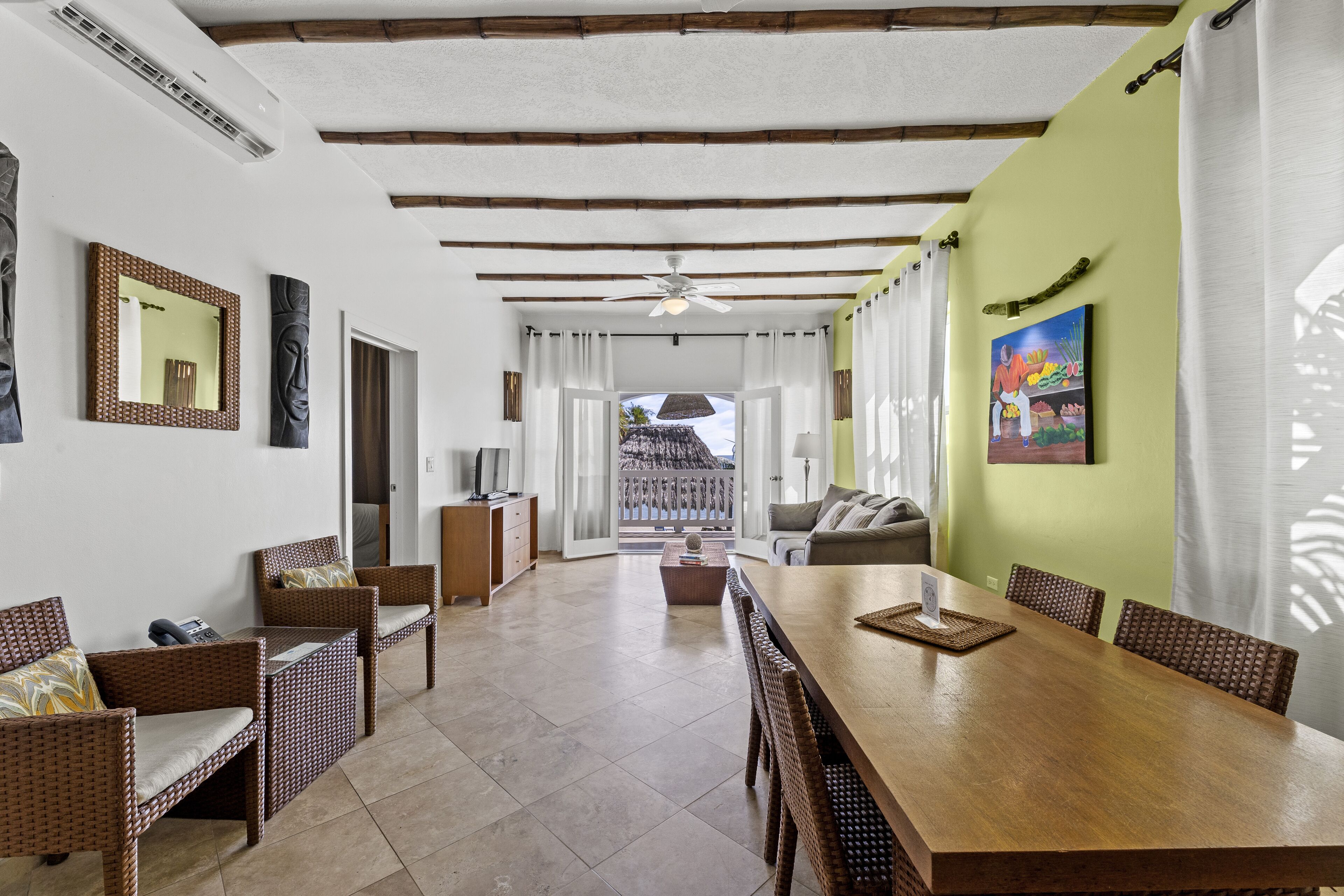 A spacious dining area with a wooden table and wicker chairs, a sofa, and a view of a thatched-roof structure through an open doorway.