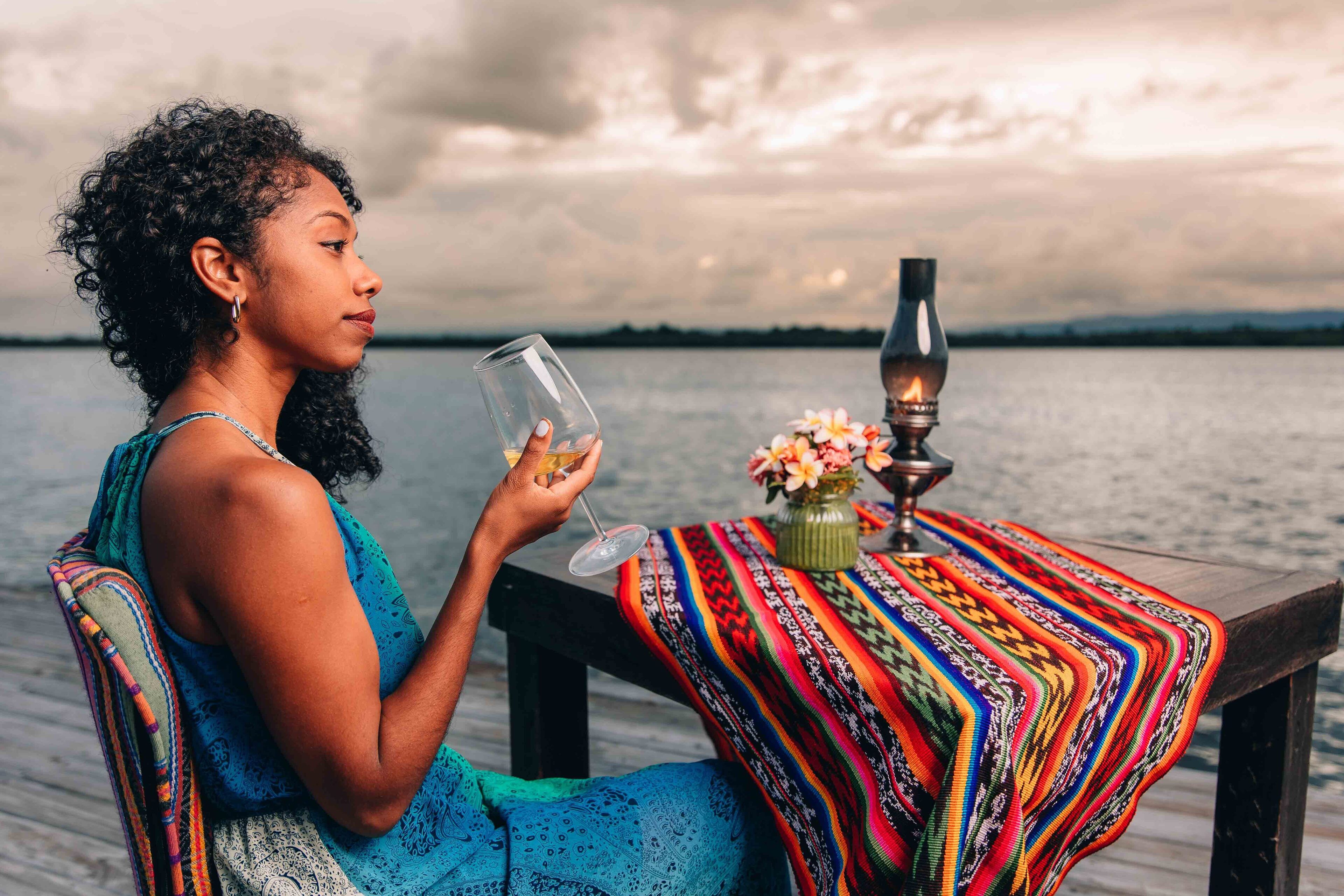 A person sitting at a wooden table by a body of water, holding a glass.