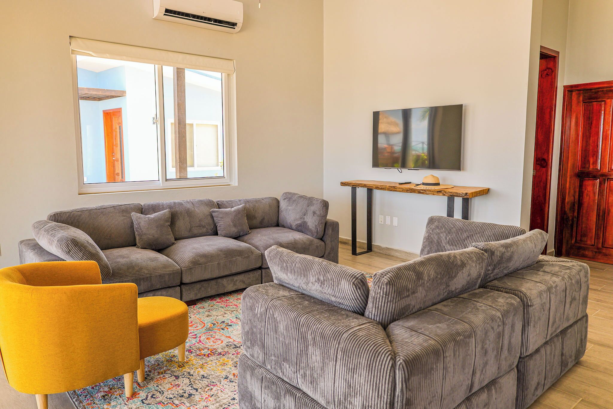 Una sala de estar con un sofá modular, un sillón amarillo, un televisor sobre un mueble de madera y una puerta roja.
