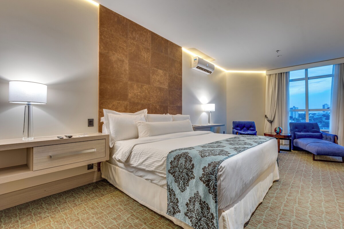 A hotel room with a large bed, bedside lamps, a desk, a chair, and a view of the city through the window.