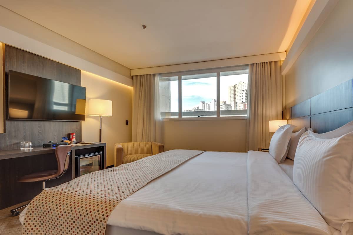 Habitación de hotel con una cama grande, un escritorio, una silla, un televisor y una ventana con vistas a la ciudad.