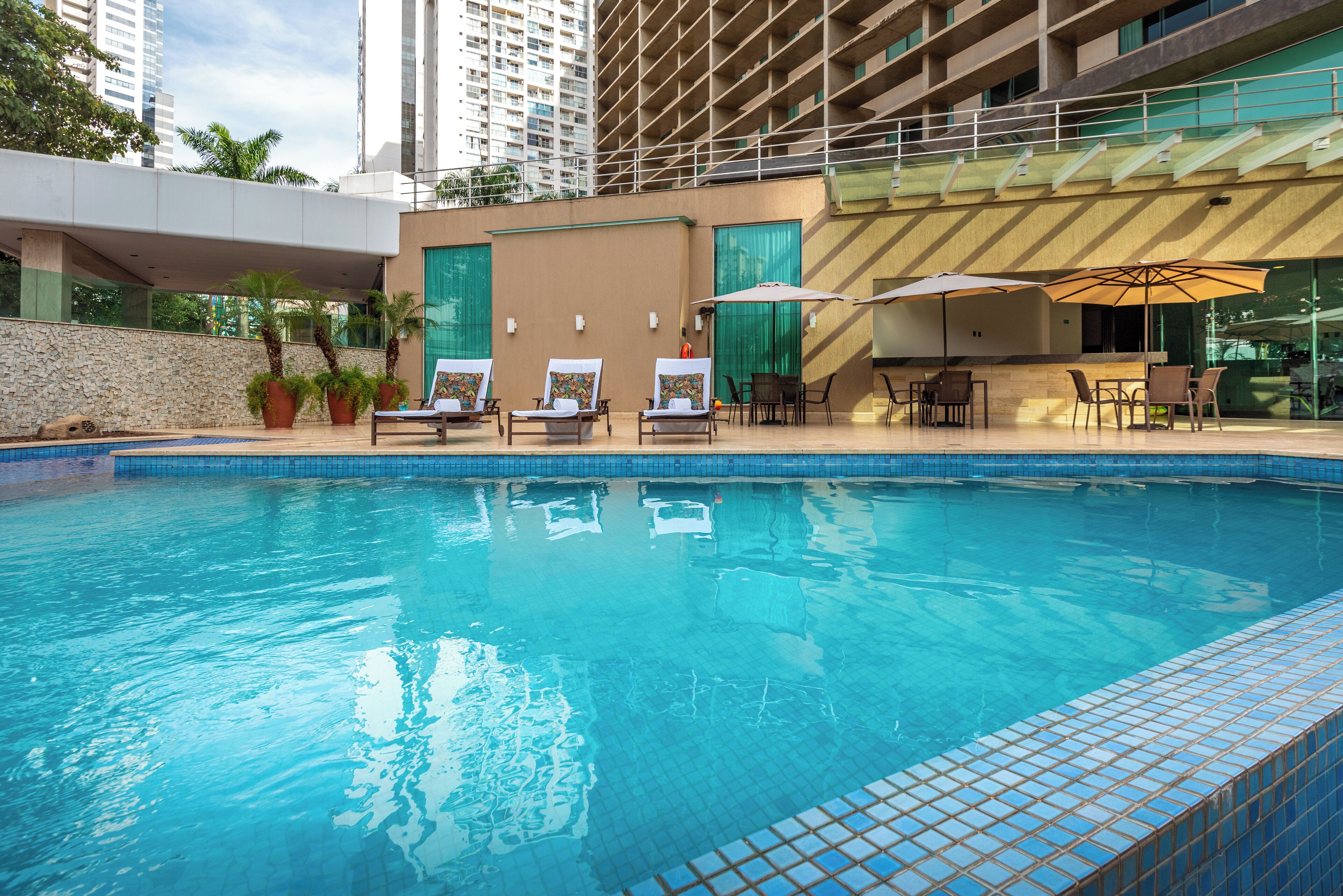 A swimming pool with lounge chairs and umbrellas, surrounded by a stone wall and modern buildings.