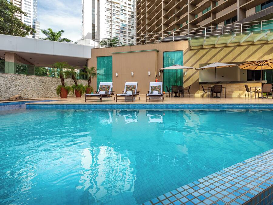 A swimming pool with lounge chairs and umbrellas, surrounded by a stone wall and modern buildings.