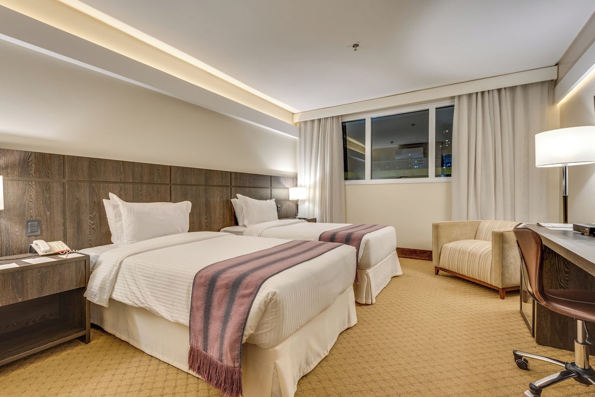 Habitación de hotel con dos camas, un escritorio, una silla, una mesita de noche con teléfono y una ventana con cortinas.