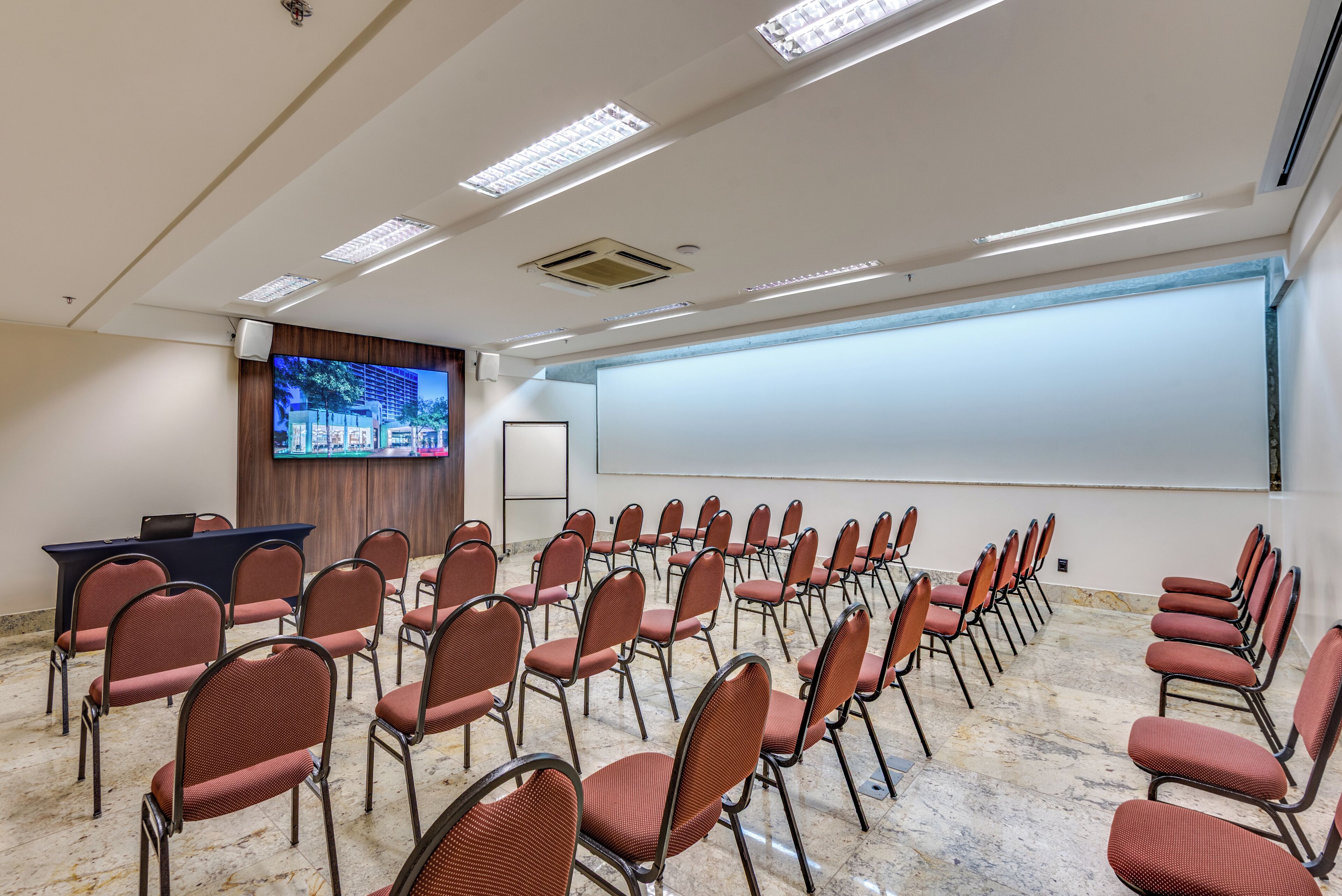 Un salón de conferencias con filas de sillas, una pantalla de proyección y un televisor.