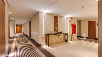 A hotel corridor with a wooden floor, a reception desk, and a red emergency exit door.