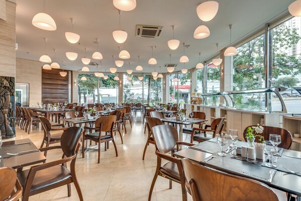 A spacious restaurant with multiple tables and chairs, large windows, and hanging pendant lights. - K Hotel (Goiania)