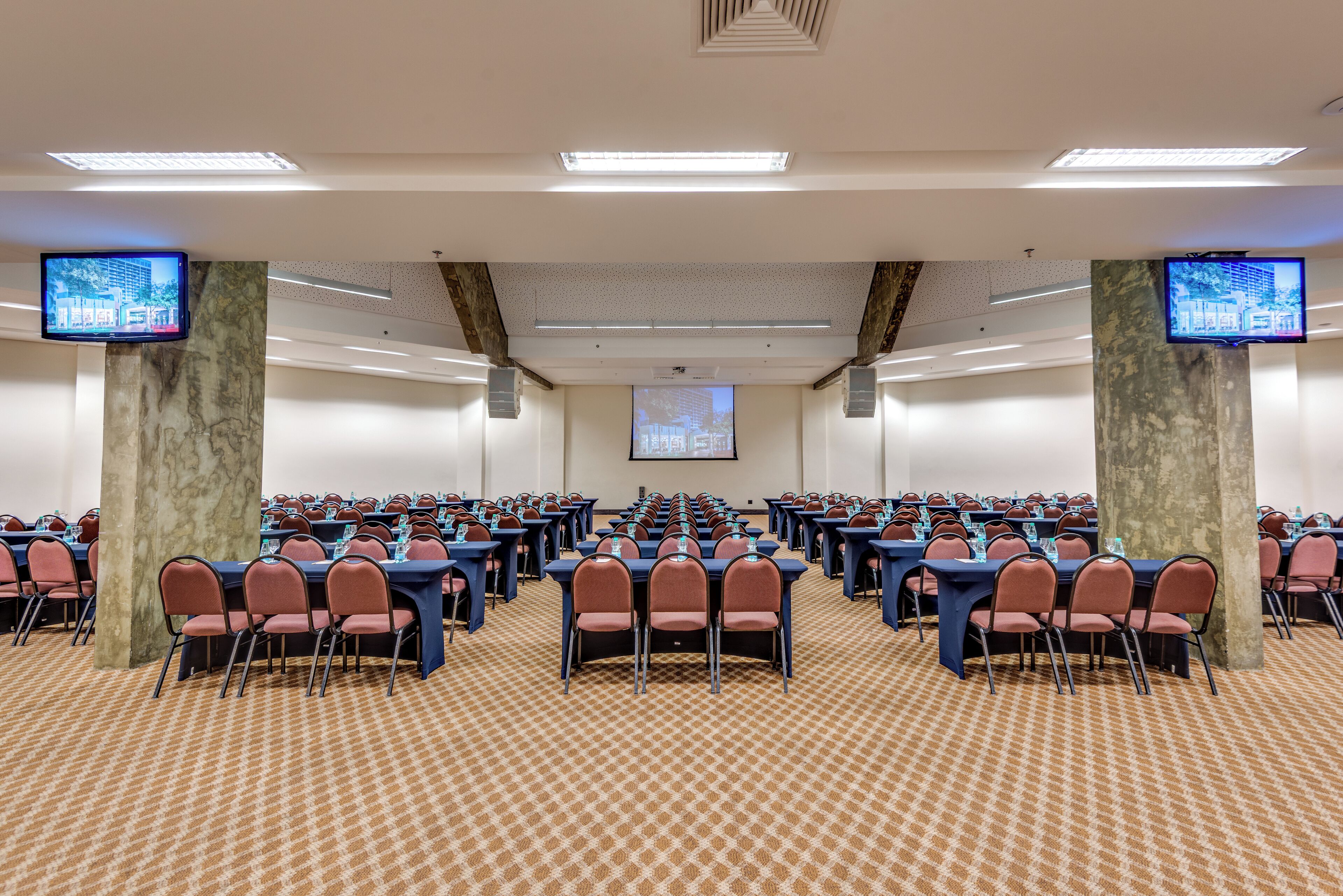 A conference room with rows of tables and chairs, equipped with water bottles and projectors, and large screens on the walls.