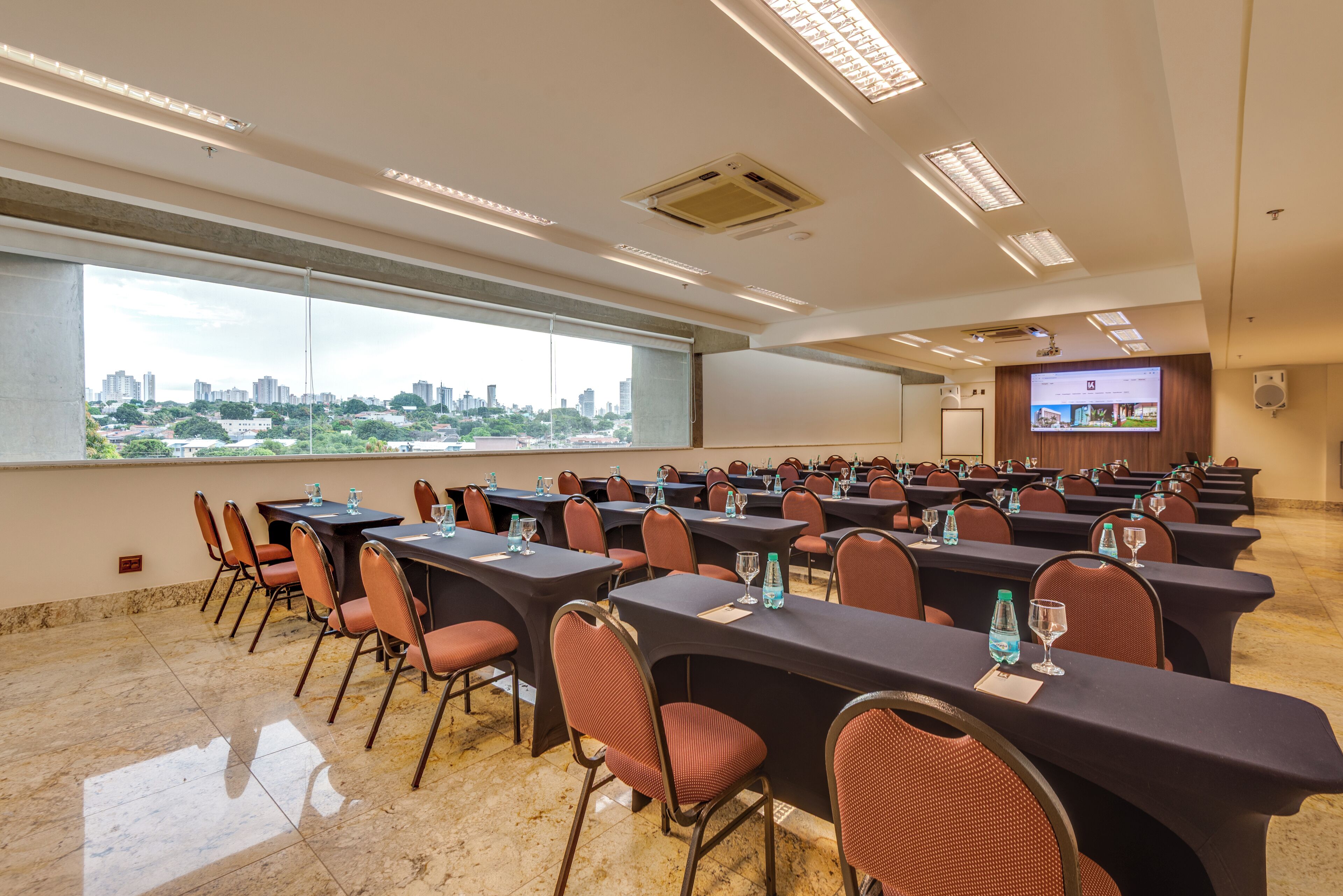 A conference room with tables and chairs, large windows with a city view, and a projection screen.