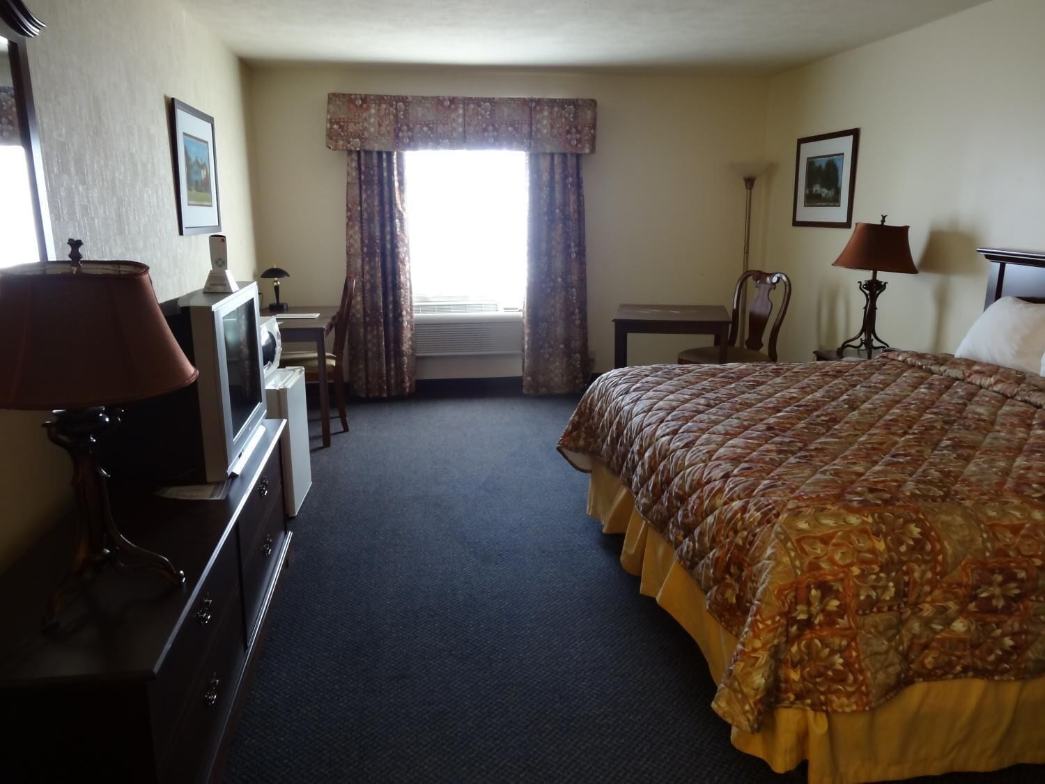 A hotel room with a bed, a desk, a chair, a television, and a lamp.