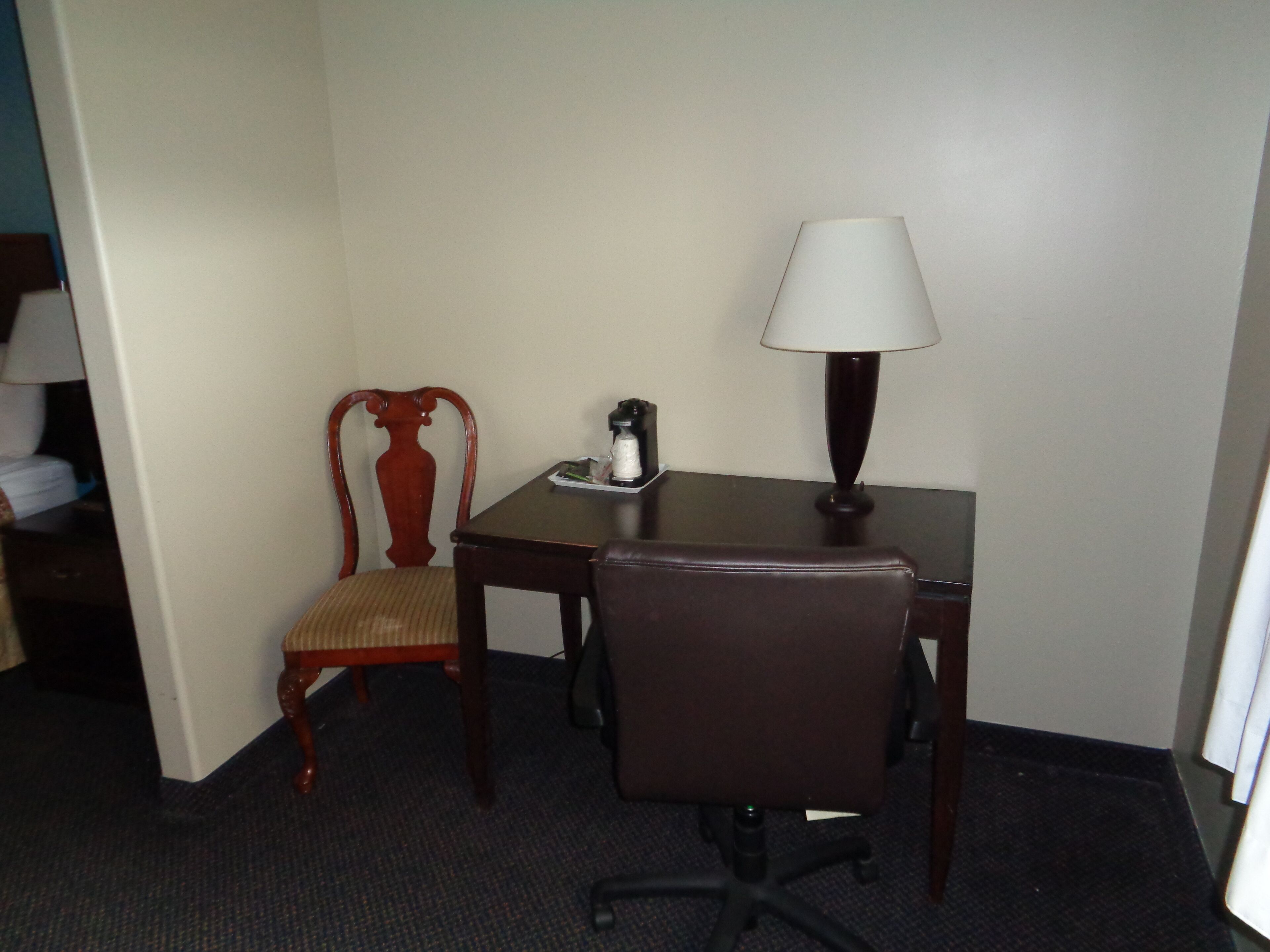 A hotel room with a wooden chair, a desk, a lamp, and a small table with a coffee maker.