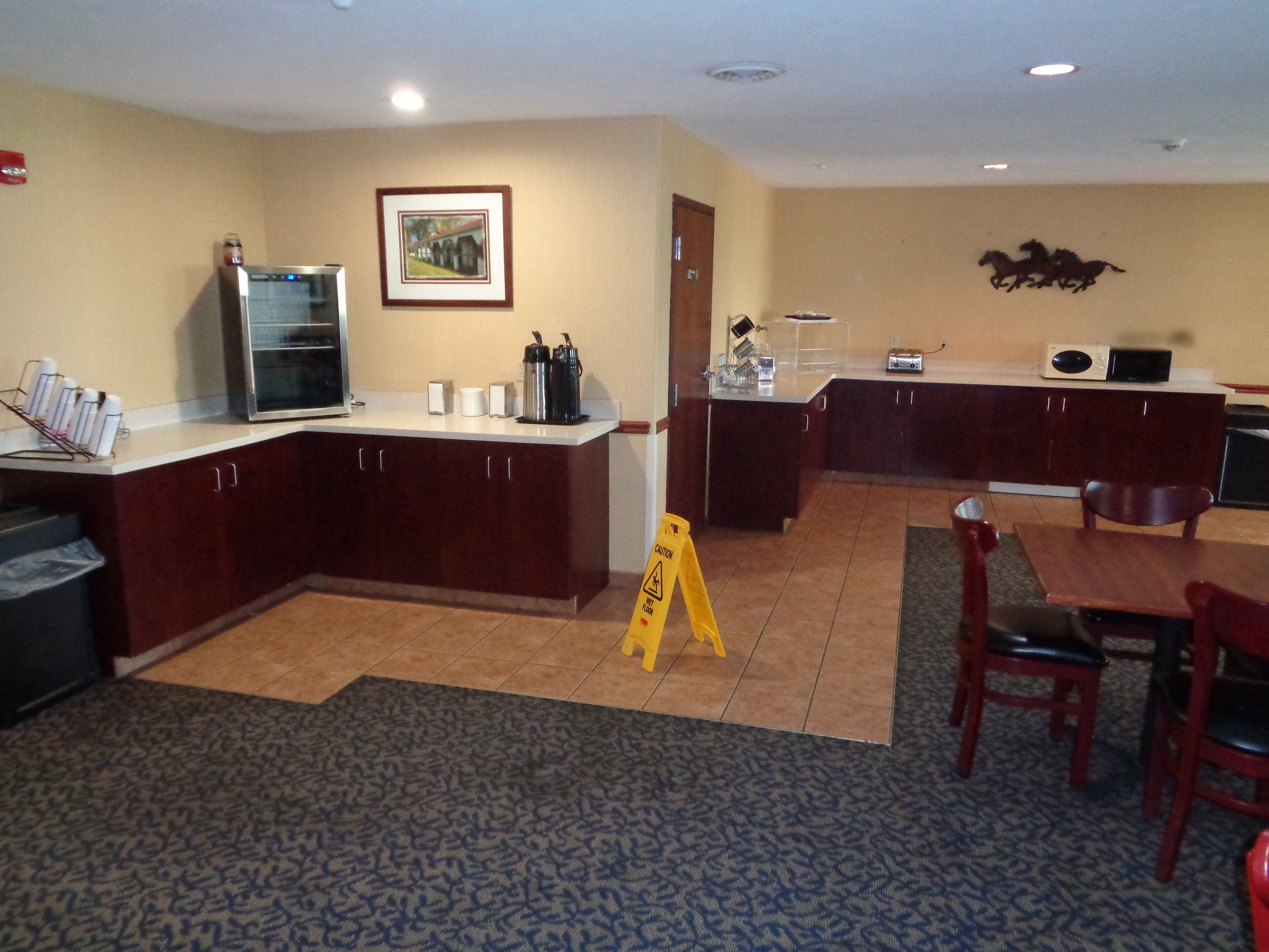 A hotel kitchen with a refrigerator, microwave, and coffee maker. There is a caution sign on the floor and a dining area with chairs and a table.