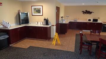 A hotel kitchen with a refrigerator, microwave, and coffee maker. There is a caution sign on the floor and a dining area with chairs and a table.