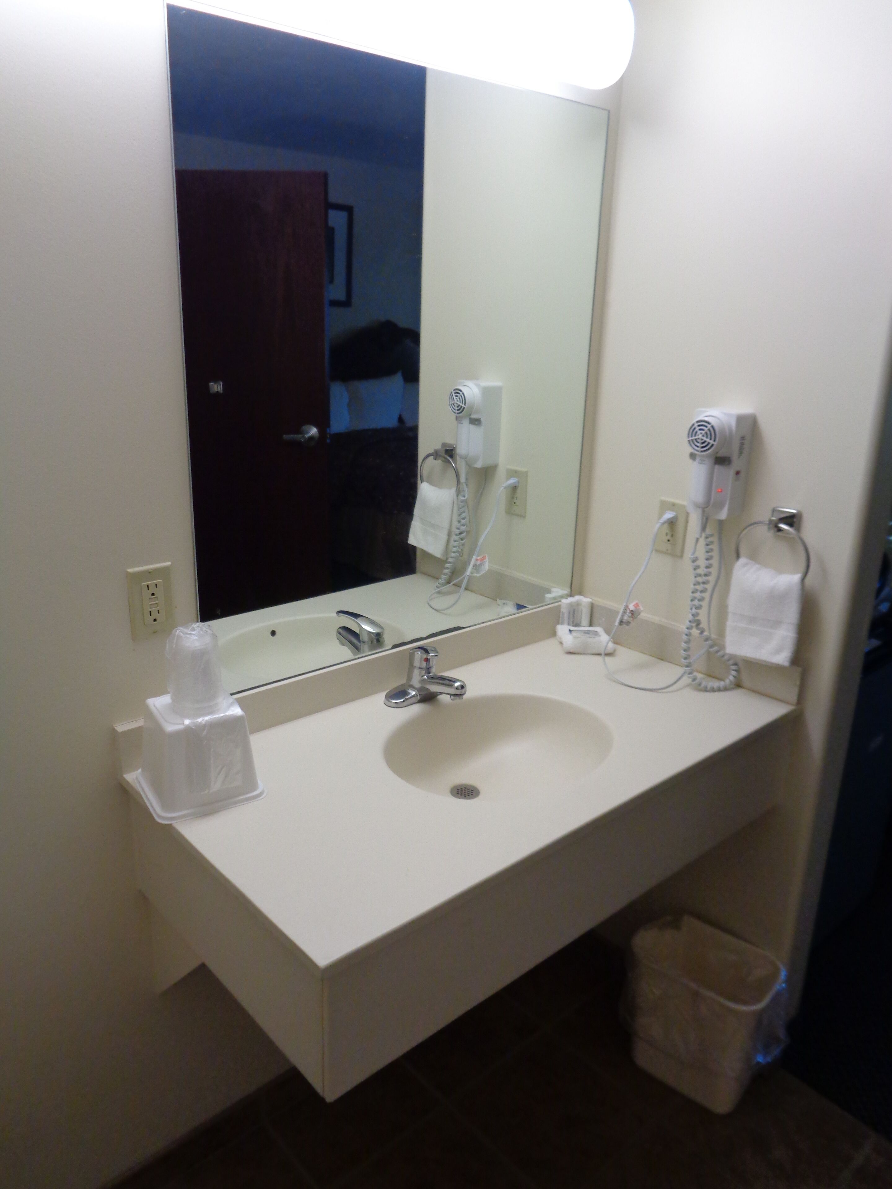 Une salle de bain d’hôtel avec un grand miroir, un lavabo, un sèche-cheveux et une poubelle.