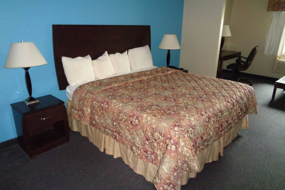 A neatly made bed with a patterned bedspread, two bedside lamps, a wooden nightstand, and a chair in a room with blue walls.