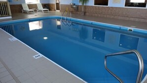 Una piscina interior con agua cristalina, una escalera metálica y sillas de descanso al fondo.