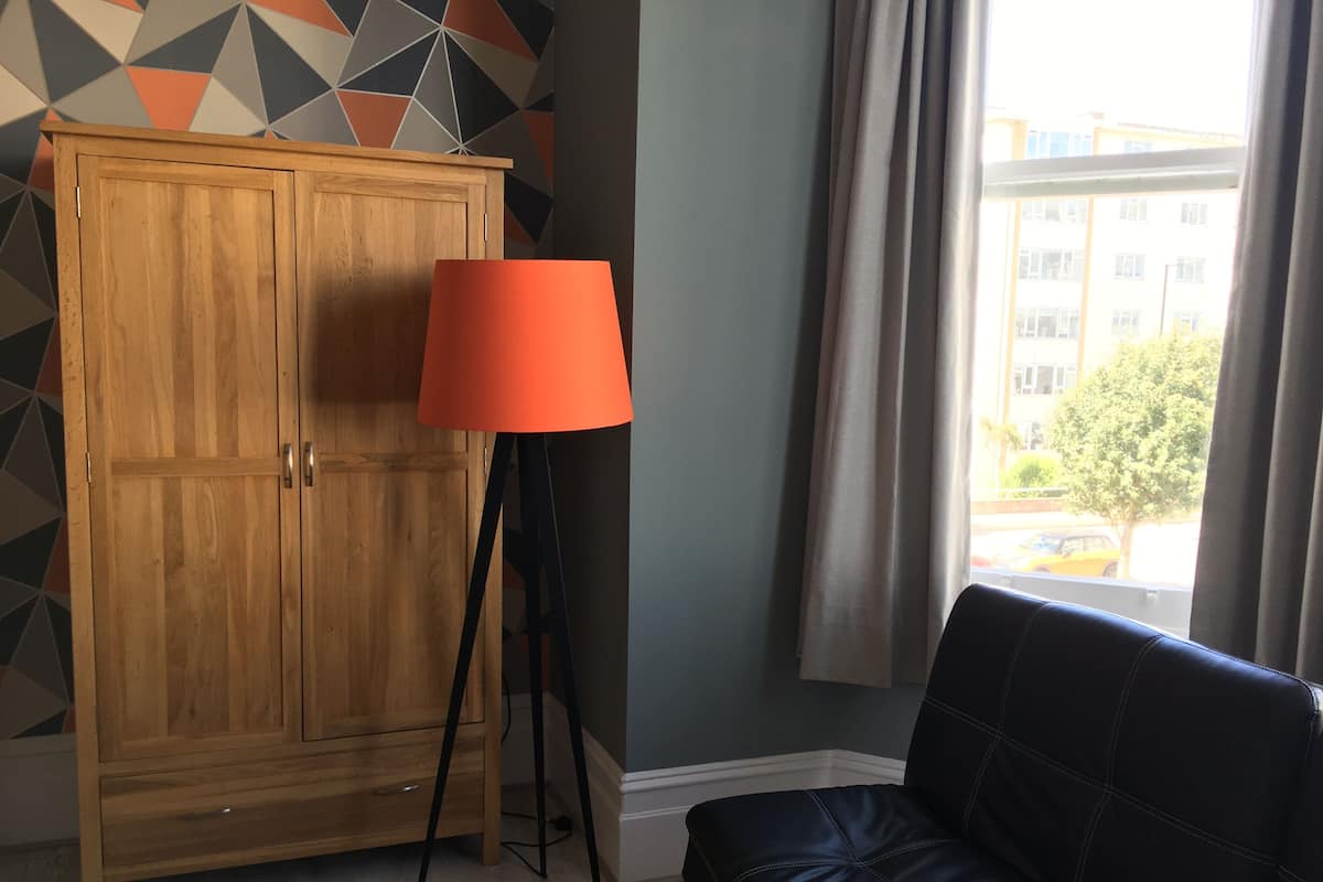 A room with a wooden wardrobe, a floor lamp, a sofa, and a window with a view of buildings and trees.