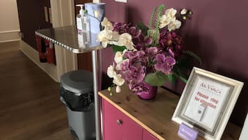 A bathroom with a purple wall, a wooden vanity, a trash can, a framed sign, and a vase of flowers.