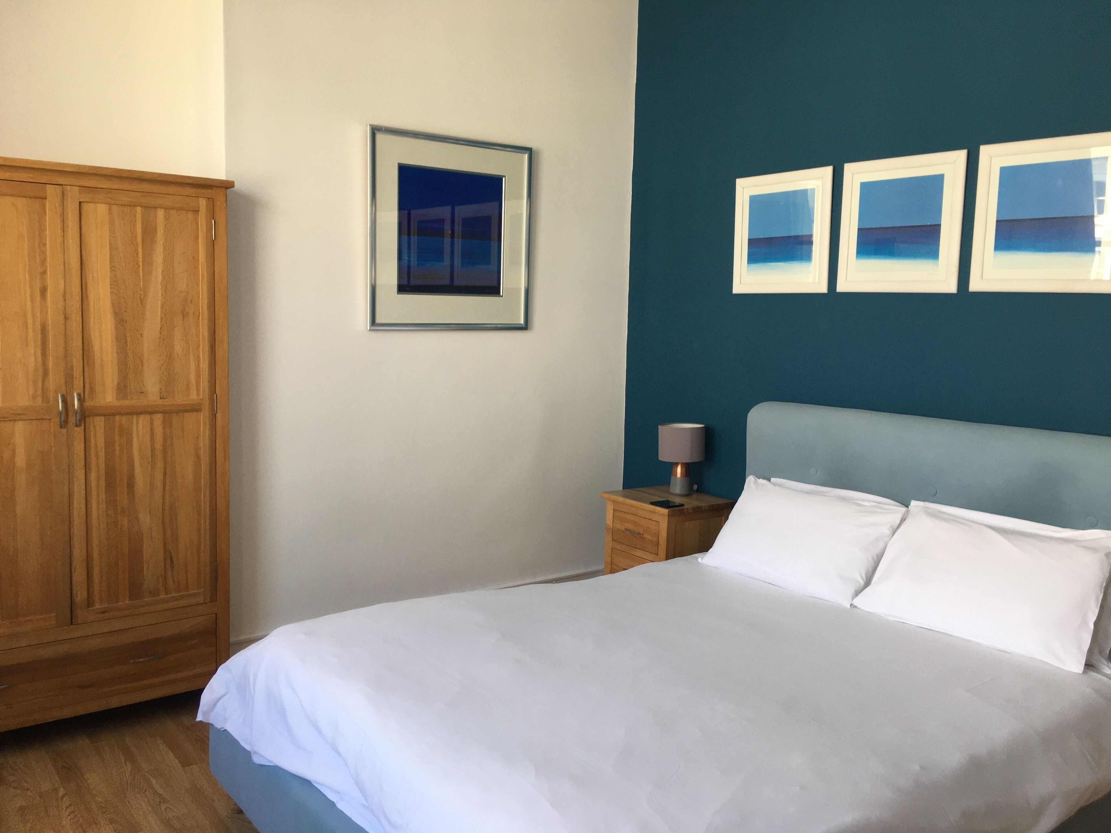 Un dormitorio con una cama grande, un armario de madera, una mesita de noche con lámpara y tres cuadros enmarcados en la pared.