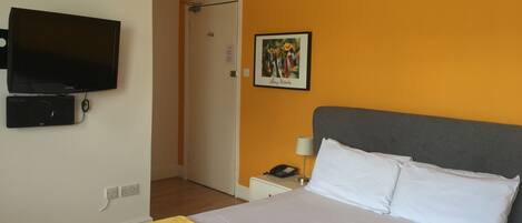 A hotel room with a bed, a TV, a bedside table with a lamp, and a door.