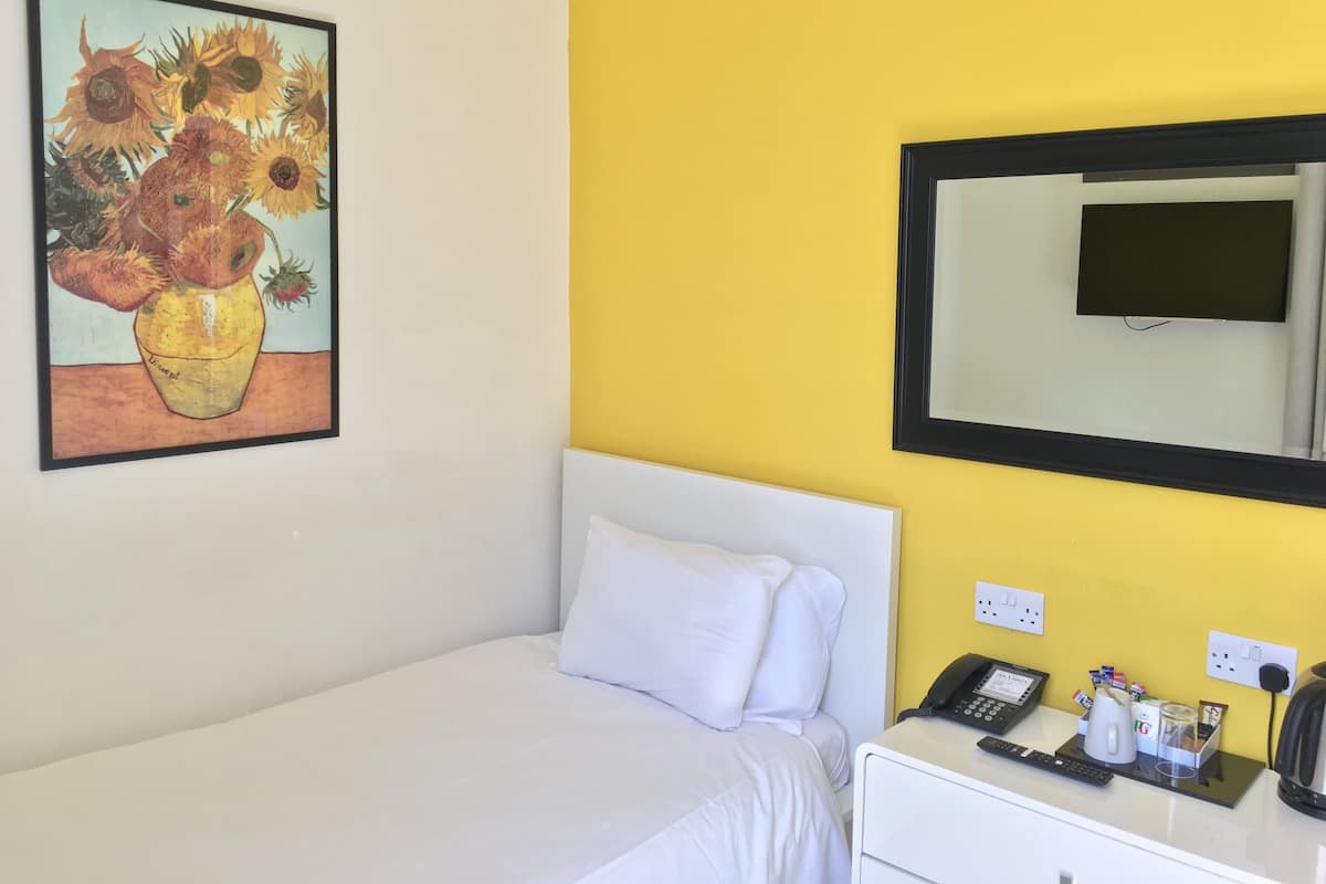 A hotel room with a bed, a bedside table with a phone and a coffee maker, a painting of sunflowers, and a mirror on the wall.