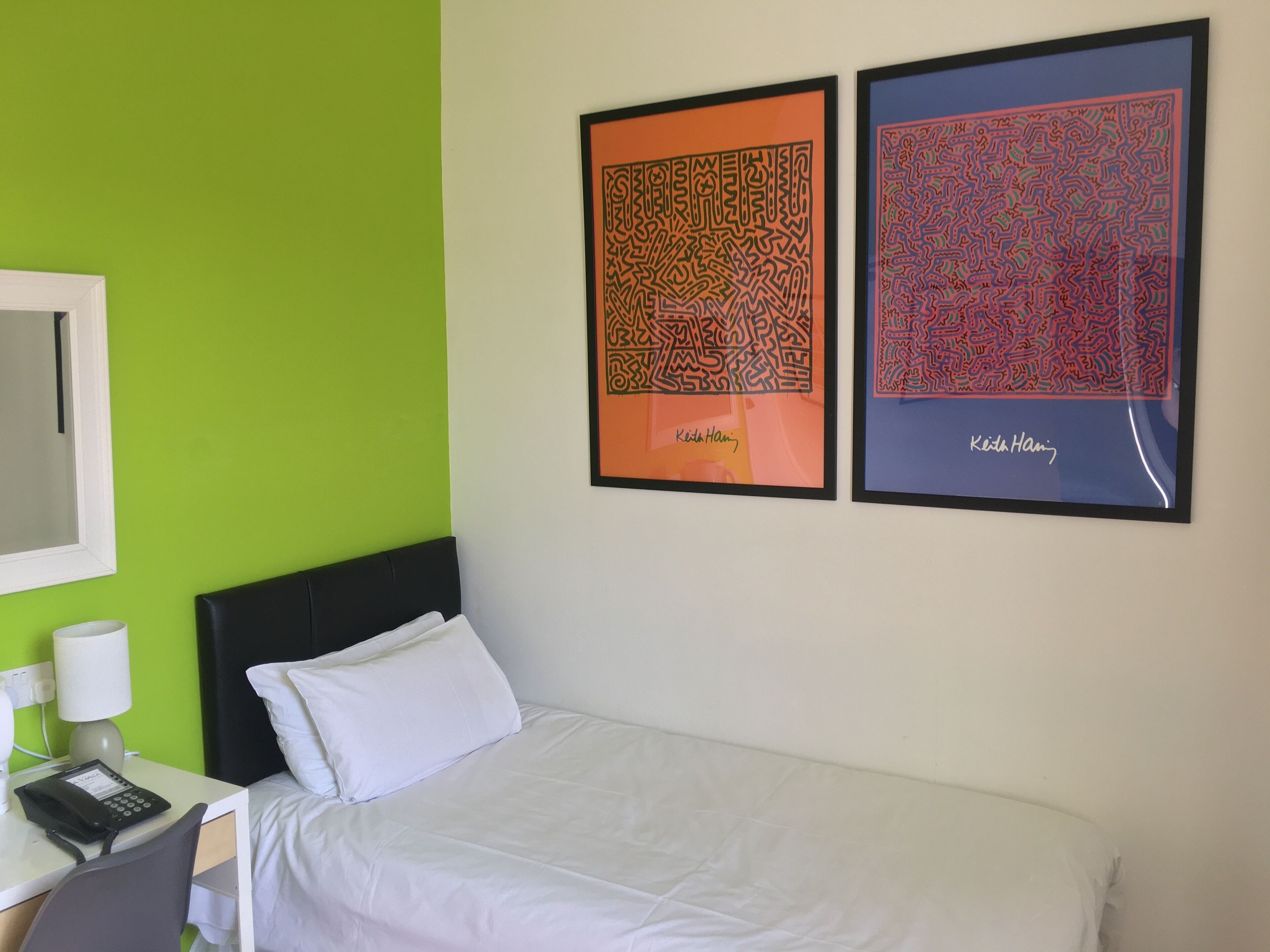 Quarto de hotel com uma cama, uma escrivaninha com telefone e duas obras de arte emolduradas na parede.
