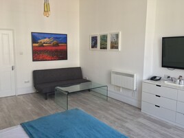 Un dormitorio moderno con un sofá gris, una mesa de centro de cristal, una alfombra azul, un aparador blanco, un televisor montado y tres cuadros en la pared.