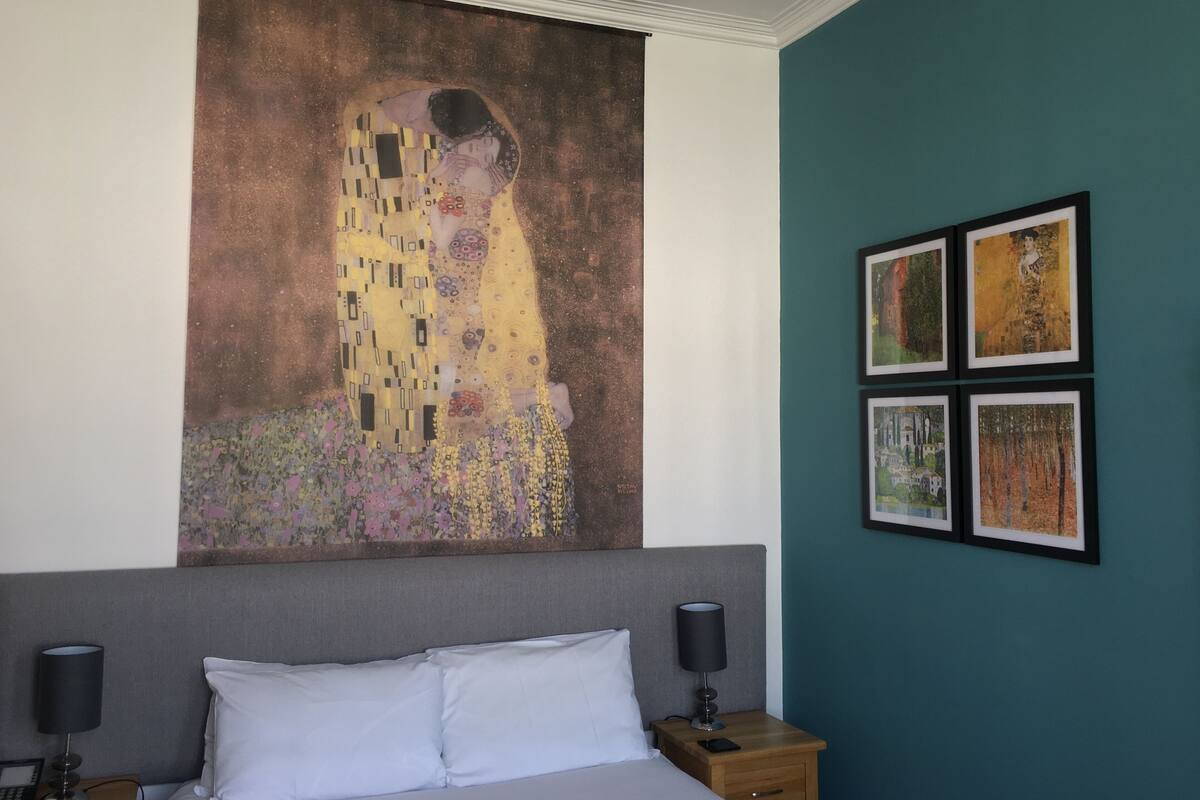 A hotel room with a large Gustav Klimt painting, a bed with white linens, a grey headboard, a nightstand with a lamp, and a teal accent wall.