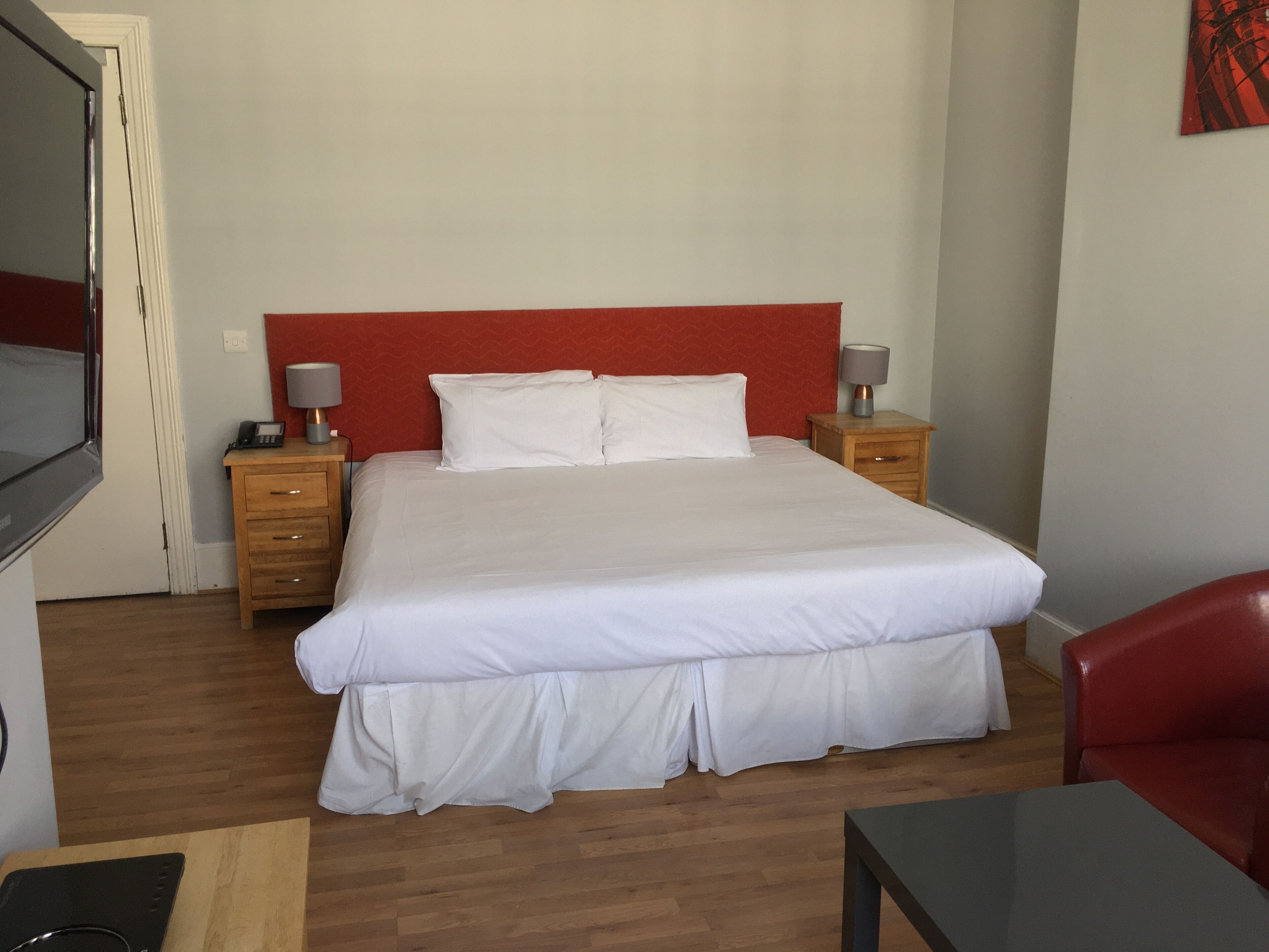 Ein Hotelzimmer mit einem großen Bett, zwei Nachttischen mit Lampen, einem Holzbeistelltisch, einem Fernseher und einem roten Ledersessel.