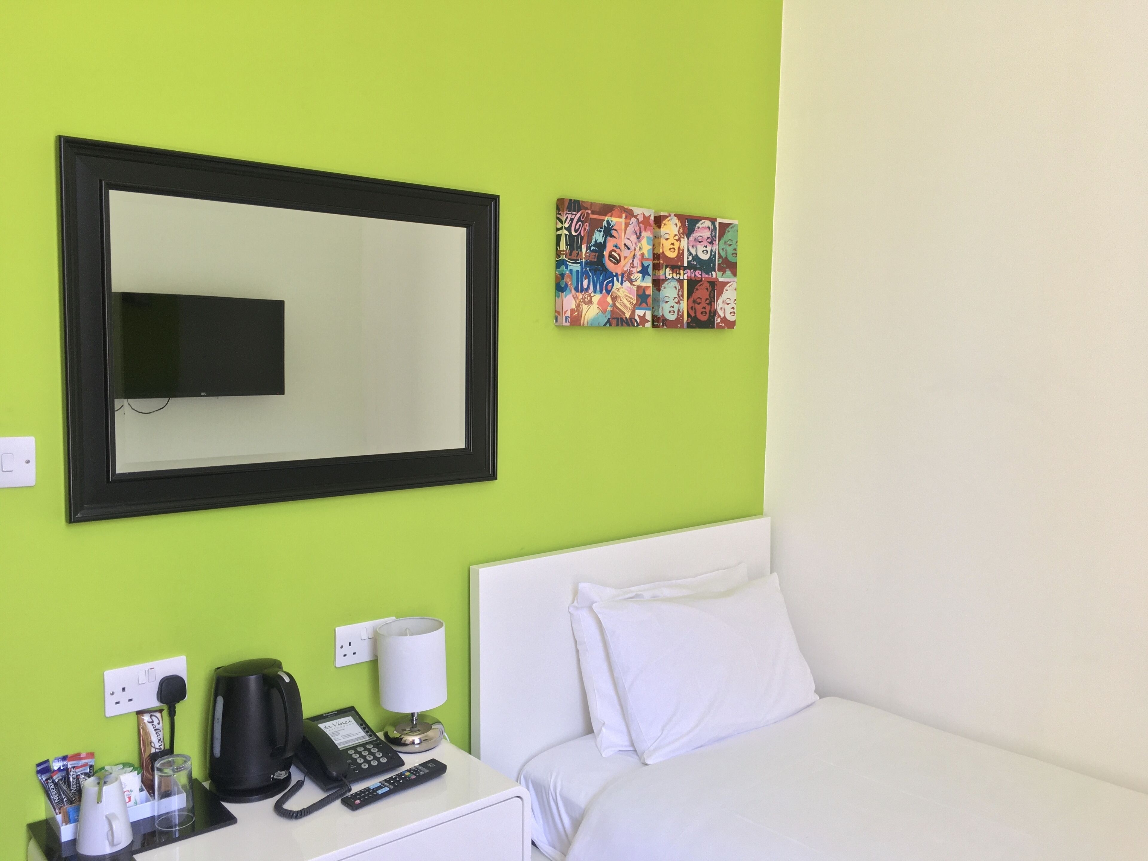Een hotelkamer met een groene wand, een grote spiegel, een bed met witte lakens, een nachtkastje met een lamp, een telefoon en een koffiezetapparaat.